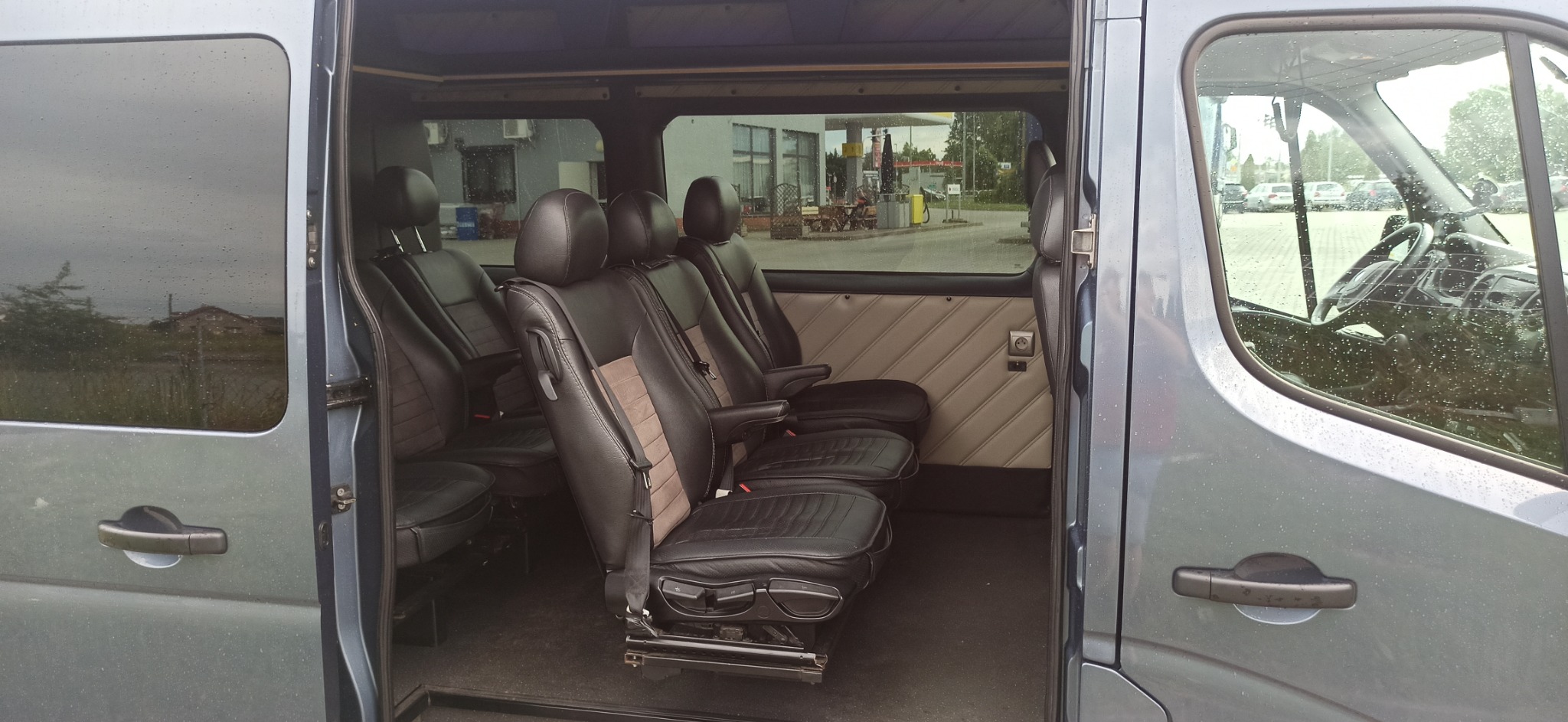 Wnętrze busa z trzema rzędami foteli, czarno-beżowa tapicerka, widok przez otwarte drzwi na zewnątrz w deszczowy dzień.