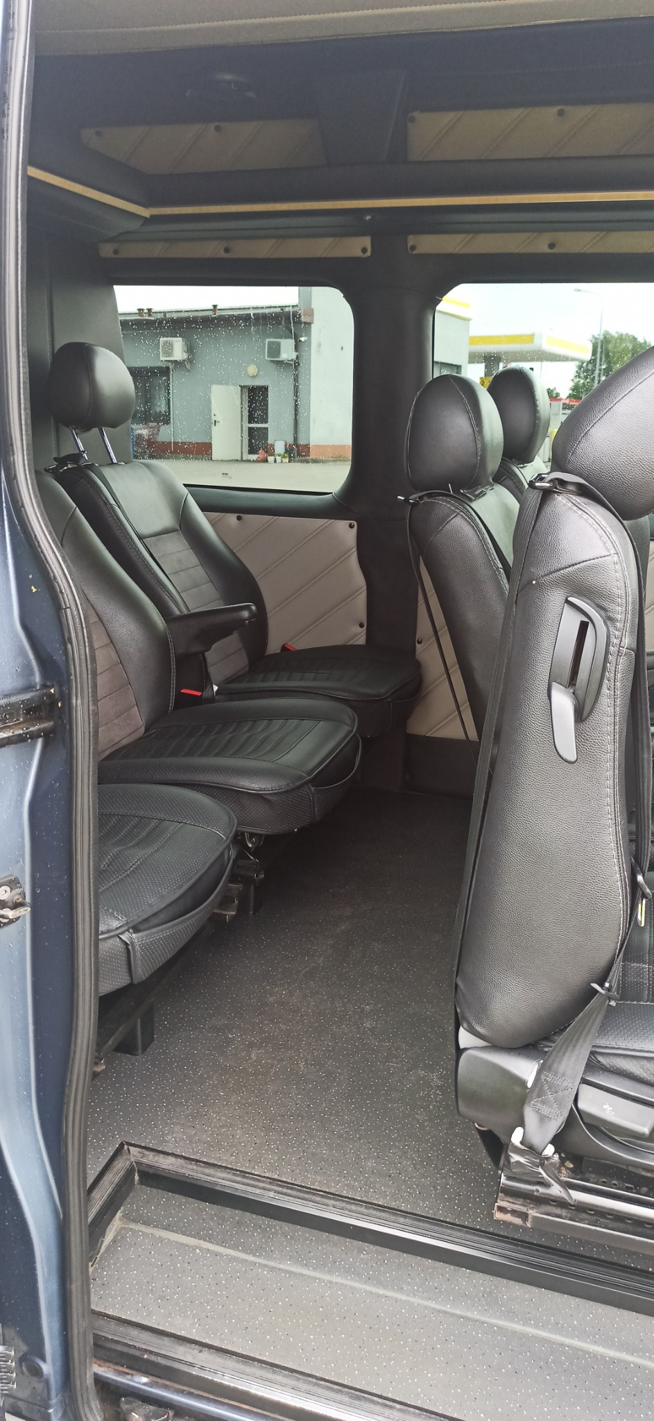 Wnętrze busa z czarnymi skórzanymi siedzeniami, widok z otwartych drzwi na trzy rzędy siedzeń, za oknem budynek stacji benzynowej w deszczowy dzień.