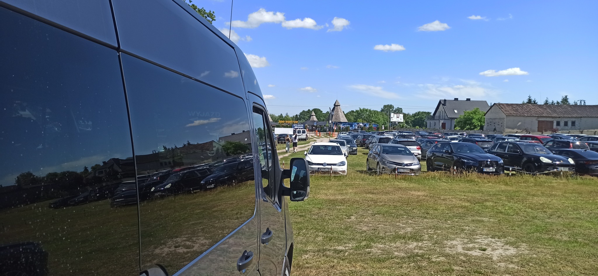 Ciemny bus na parkingu, w odbiciu na szybie widoczny budynek i zaparkowane samochody, w tle tablica informacyjna Zoo Borysew.