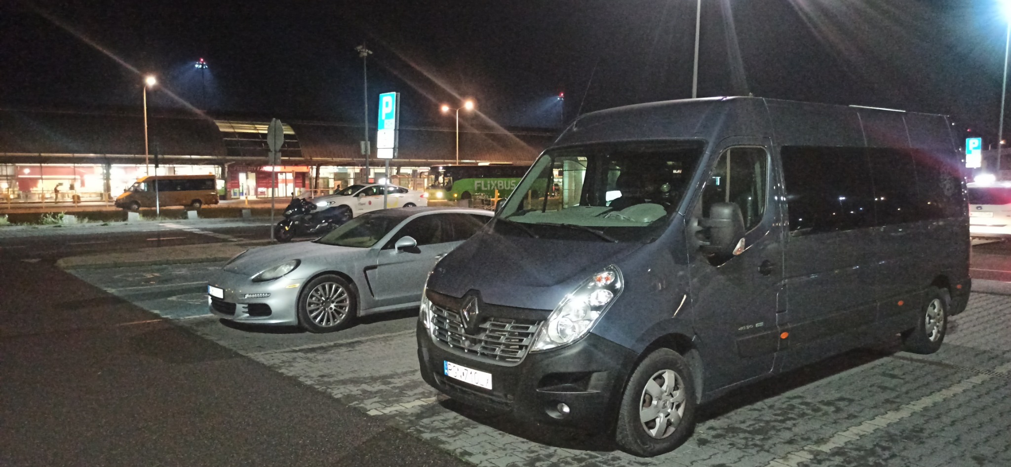 Ciemny bus Renault Master na parkingu lotniska nocą, obok srebrny Porsche Panamera, w tle budynek terminala i autobus Flixbus.