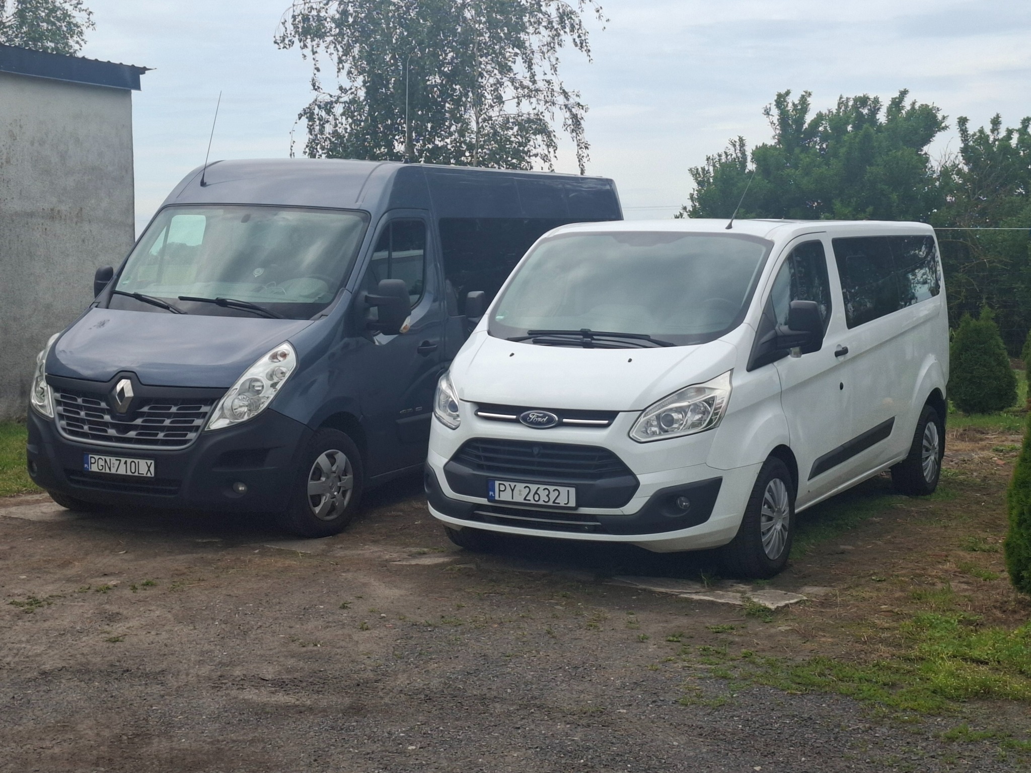 Dwa busy: biały Ford Transit i szary Renault Master, zaparkowane obok siebie na żwirowym podłożu, z zielenią w tle. Tablice rejestracyjne widoczne.