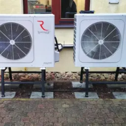 Dwie białe pompy ciepła Rotenso Aquami, zamontowane na czarnych metalowych stelażach, ustawione na brukowanym podłożu przed jasną ścianą budynku z oknem.