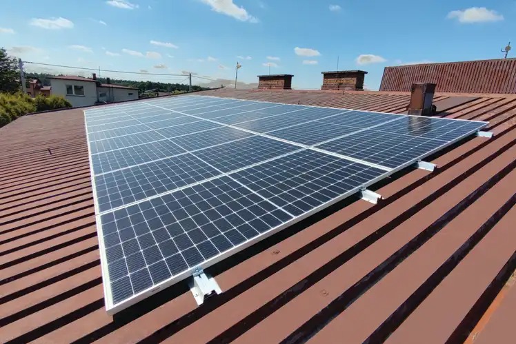 Panele słoneczne zamontowane na brązowym, blaszanym dachu budynku jednorodzinnego, widoczne kominy i błękitne niebo z chmurami.