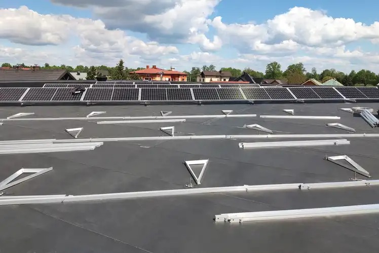 Montaż paneli słonecznych na dachu: widoczne panele w górnej części kadru oraz aluminiowe konstrukcje wsporcze w dolnej części, czekające na montaż kolejnych paneli. W tle domy mieszkalne...