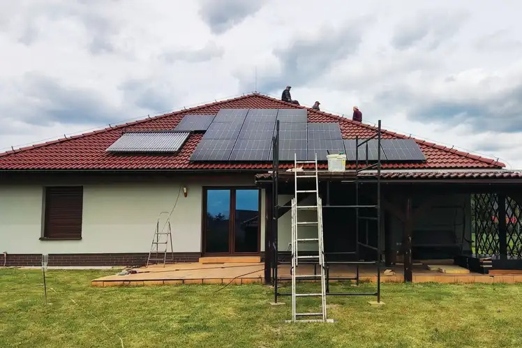 Montaż paneli słonecznych i kolektora słonecznego na dachu domu jednorodzinnego z czerwoną dachówką, widoczni pracownicy na dachu, drabina i rusztowanie przy elewacji.