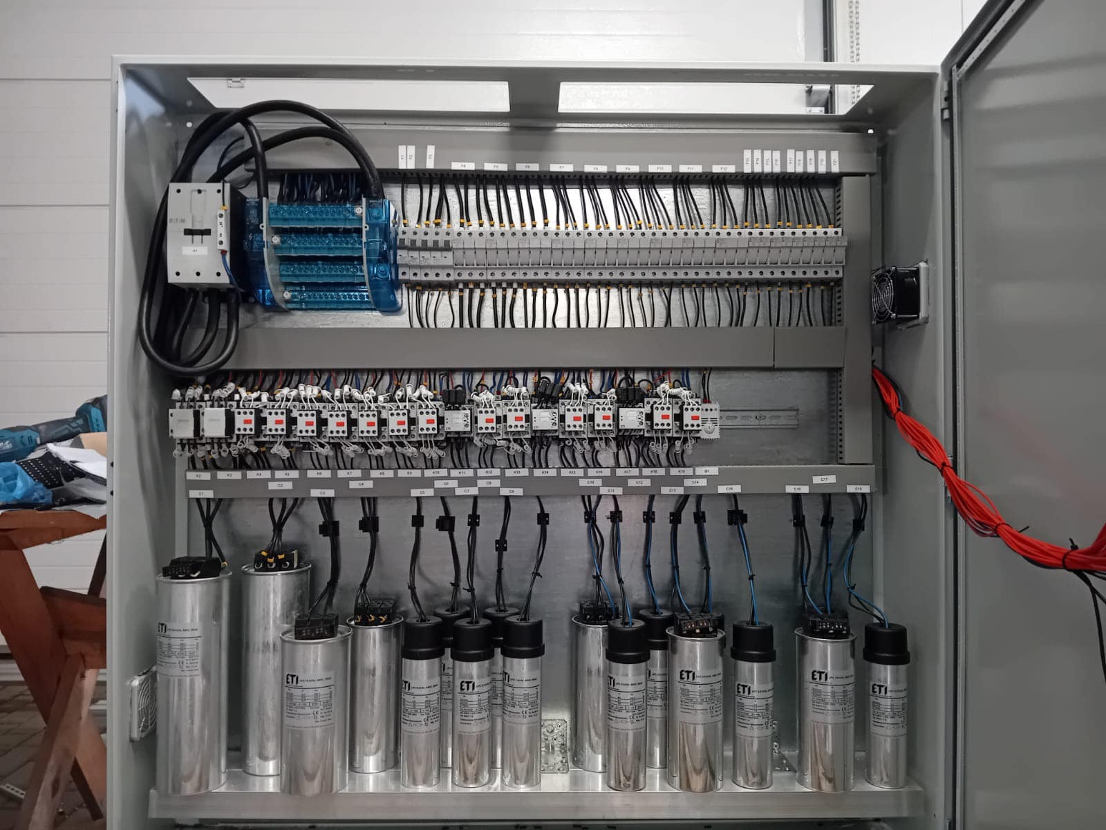Otwarta szafa sterownicza z kondensatorami ETI, licznymi przewodami i listwami zaciskowymi, wentylatorem chłodzącym po prawej stronie i niebieskimi elementami po lewej, widoczne narzędzia...