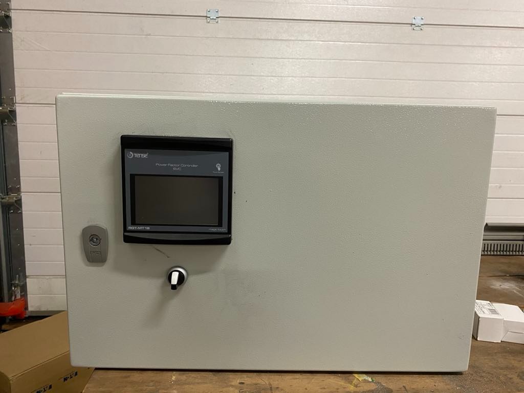 Szara szafa sterownicza z wyświetlaczem Power Factor Controller SVC firmy Tense, zamontowana na tle białej bramy garażowej.