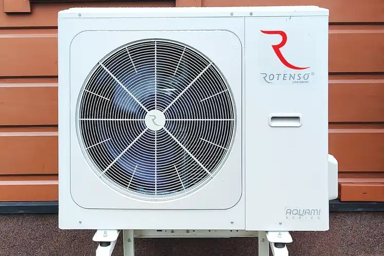 Biała jednostka zewnętrzna z wentylatorem i logo Rotenso Aquami Series zamontowana przy brązowej elewacji budynku.