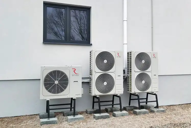 Trzy jednostki zewnętrzne z logo producenta, zamontowane na metalowych podstawach na betonowych bloczkach przy białej ścianie budynku z szarym oknem. Widoczna rura spustowa.