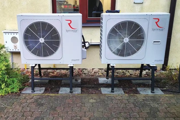 Dwie białe pompy ciepła Rotenso Aquami, zamontowane na czarnych metalowych stelażach, ustawione na brukowanym podłożu przed jasną ścianą budynku z oknem.