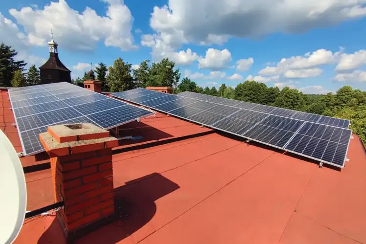Panele słoneczne na czerwonym dachu budynku z ceglanym kominem i wieżą kościoła w tle, widok w słoneczny dzień.