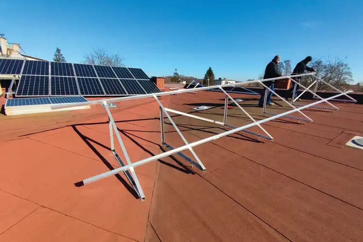 Montaż konstrukcji wsporczej pod panele słoneczne na dachu pokrytym czerwoną papą, widoczne już zamontowane panele w tle i dwóch monterów przy pracy w słoneczny dzień.