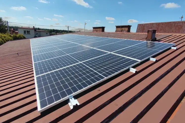Panel fotowoltaiczny zamontowany na brązowym, blaszanym dachu domu jednorodzinnego, widok z góry przy słonecznej pogodzie.