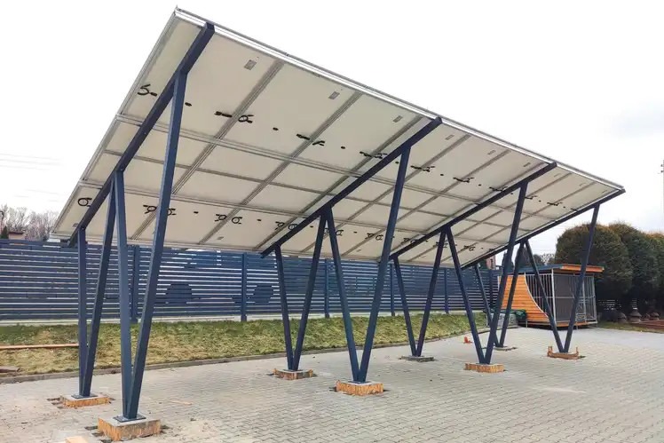 Zadaszenie parkingu z panelami słonecznymi od spodu, wsparte na metalowych, skośnych podporach, na kostce brukowej, w tle ogrodzenie i budynek.