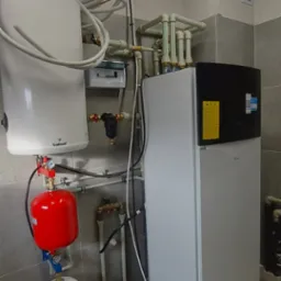 Biała pompa ciepła Daikin z czarnym panelem, podłączona do instalacji z czerwonym zbiornikiem i białym zasobnikiem, widoczna również rozdzielnica elektryczna, rury i szare płytki na ścianie...