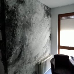 Alva Instalacje Sebastian Post - Ściana w salonie pokryta dekoracyjnym tynkiem w odcieniach szarości, przypominającym marmurkowy wzór, z widocznym grzejnikiem i fragmentem fotela.