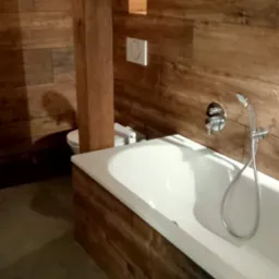 Wnętrze łazienki z drewnianą boazerią, białą wanną z chromowaną armaturą i toaletą w tle, oświetlone punktowym światłem.