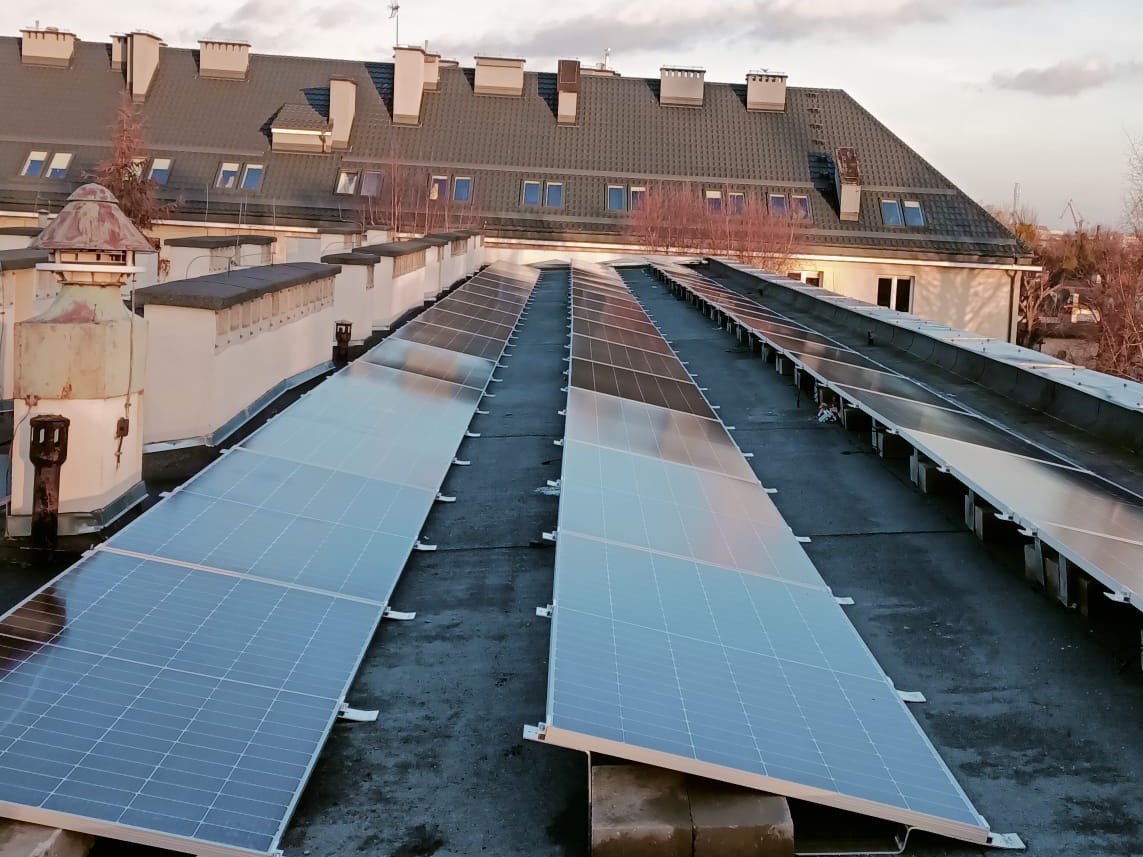 Rząd paneli fotowoltaicznych na płaskim dachu budynku mieszkalnego, widoczne metalowe mocowania, w tle kominy wentylacyjne i dachówka sąsiedniego budynku.