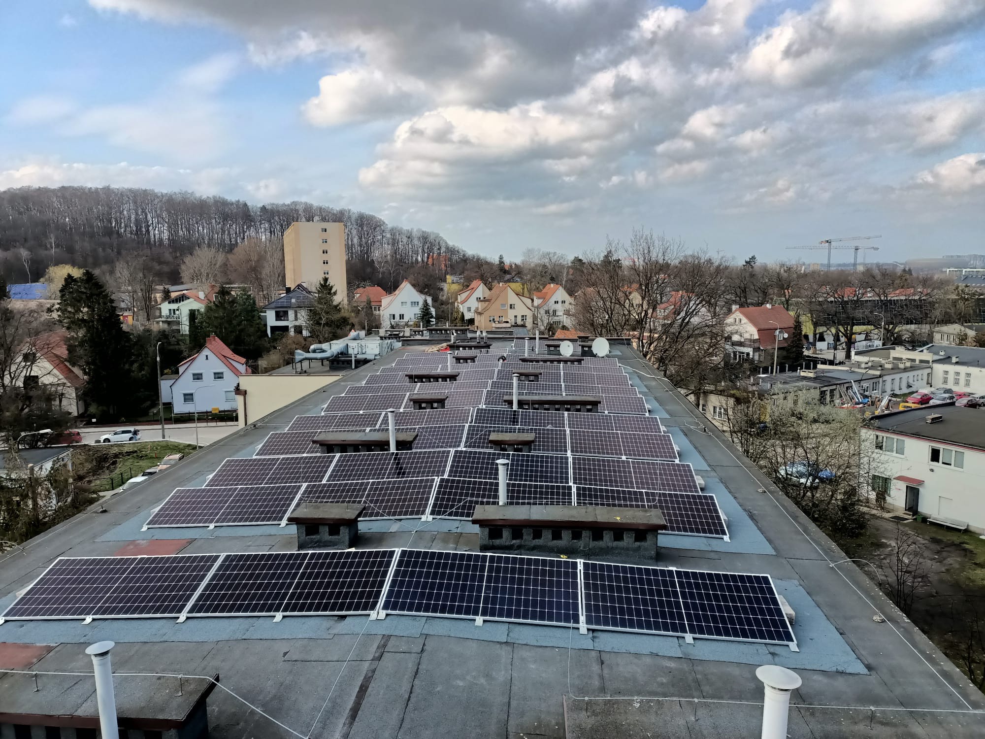 Instalacja fotowoltaiczna na płaskim dachu budynku mieszkalnego, widok z góry na panele słoneczne z kominami wentylacyjnymi i otoczeniem miejskim w tle, pochmurne niebo.
