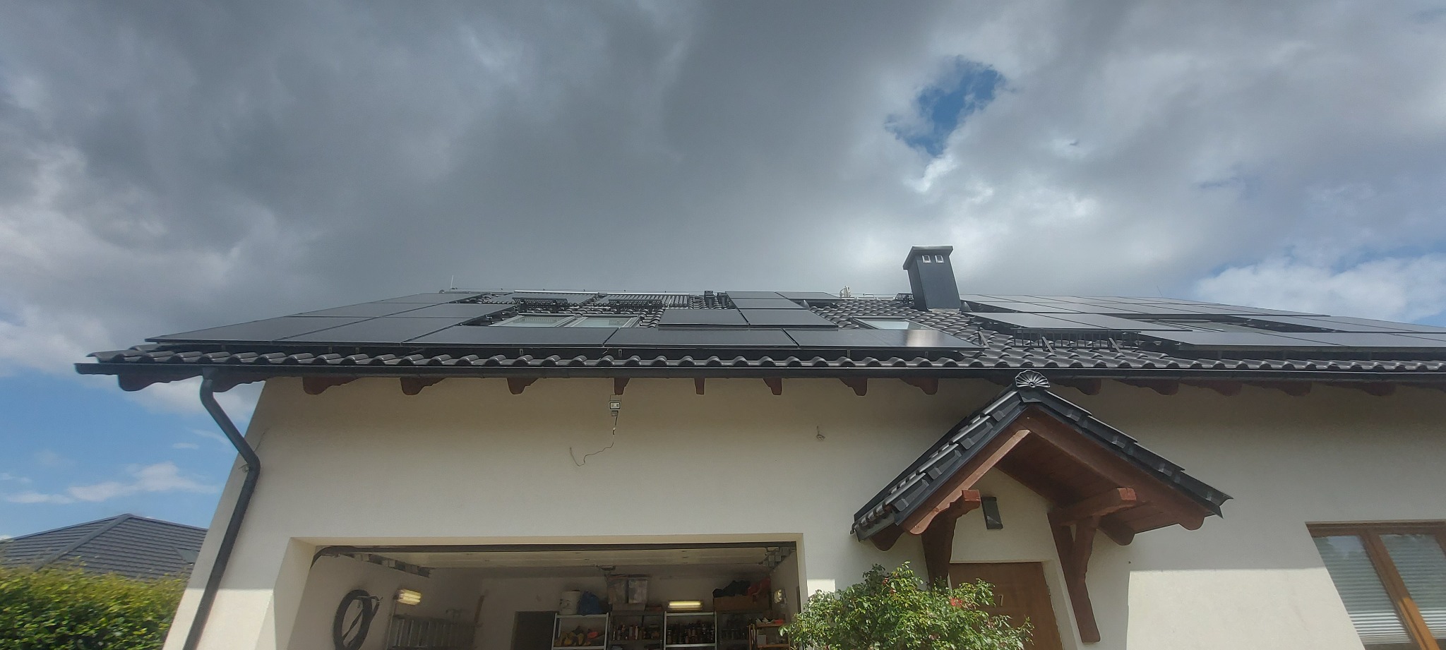 Dom jednorodzinny z zainstalowanymi panelami fotowoltaicznymi na dachu pokrytym dachówką, widoczne okna dachowe i komin, pochmurne niebo w tle.