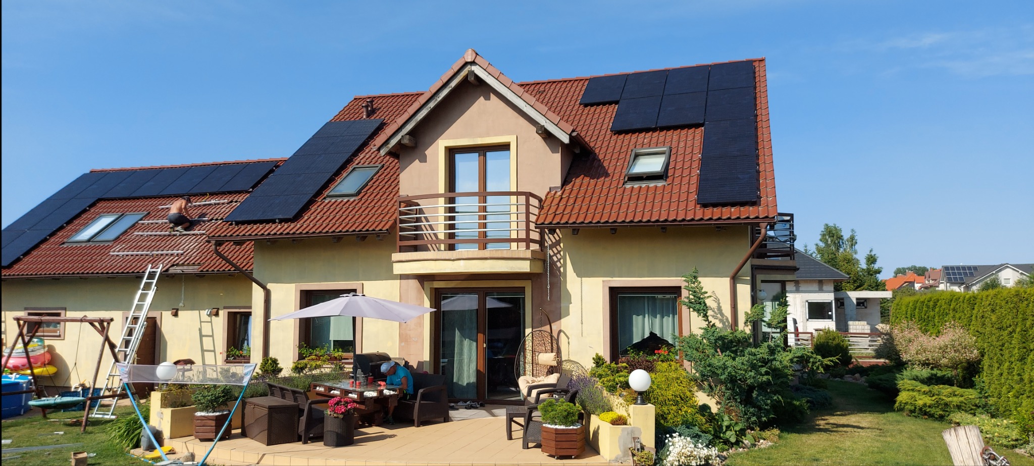 Montaż paneli fotowoltaicznych na dachu dwupiętrowego domu z czerwonej dachówki; widoczny monter na dachu, rusztowania i częściowo zainstalowane panele, taras z meblami ogrodowymi w tle.