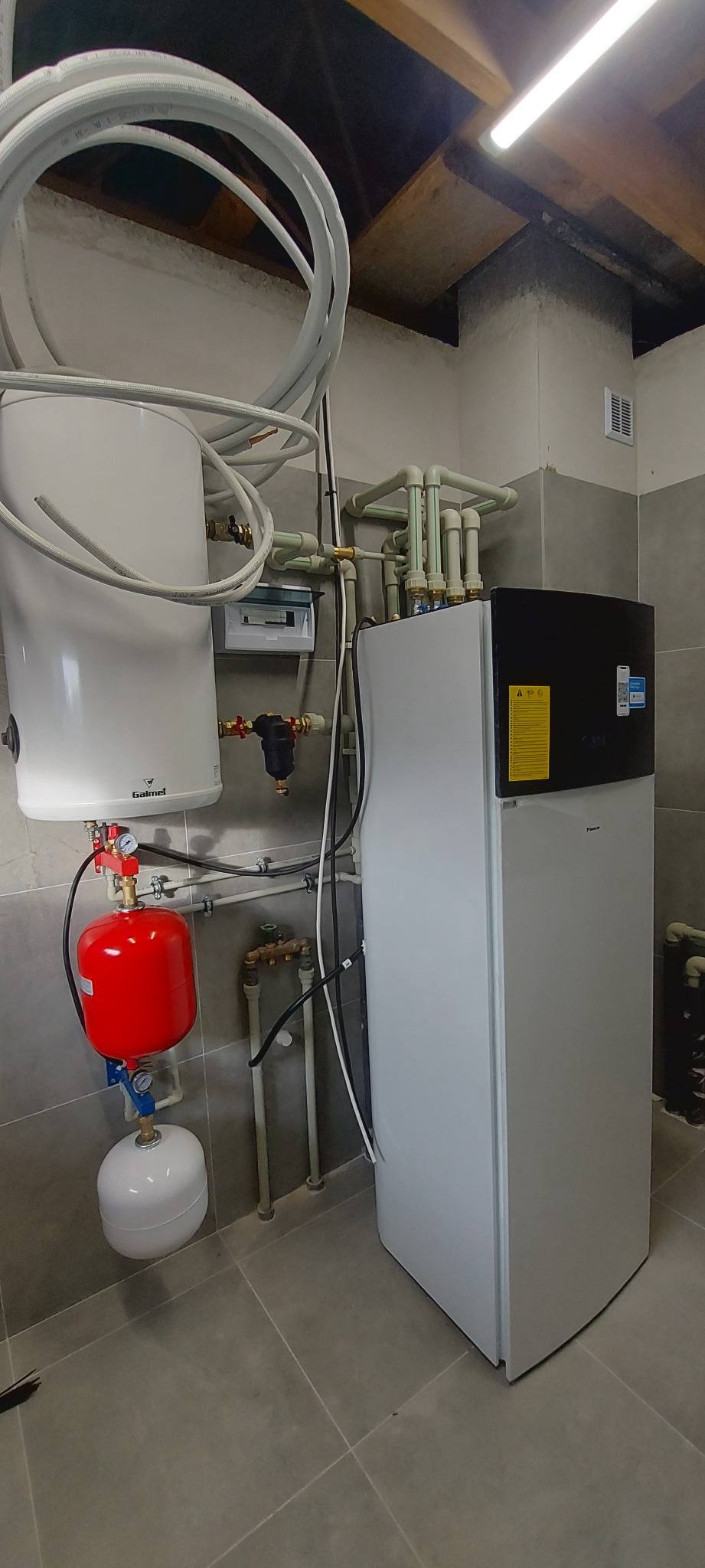 Biała pompa ciepła Daikin z czarnym panelem, podłączona do instalacji z czerwonym zbiornikiem i białym zasobnikiem, widoczna również rozdzielnica elektryczna, rury i szare płytki na ścianie...