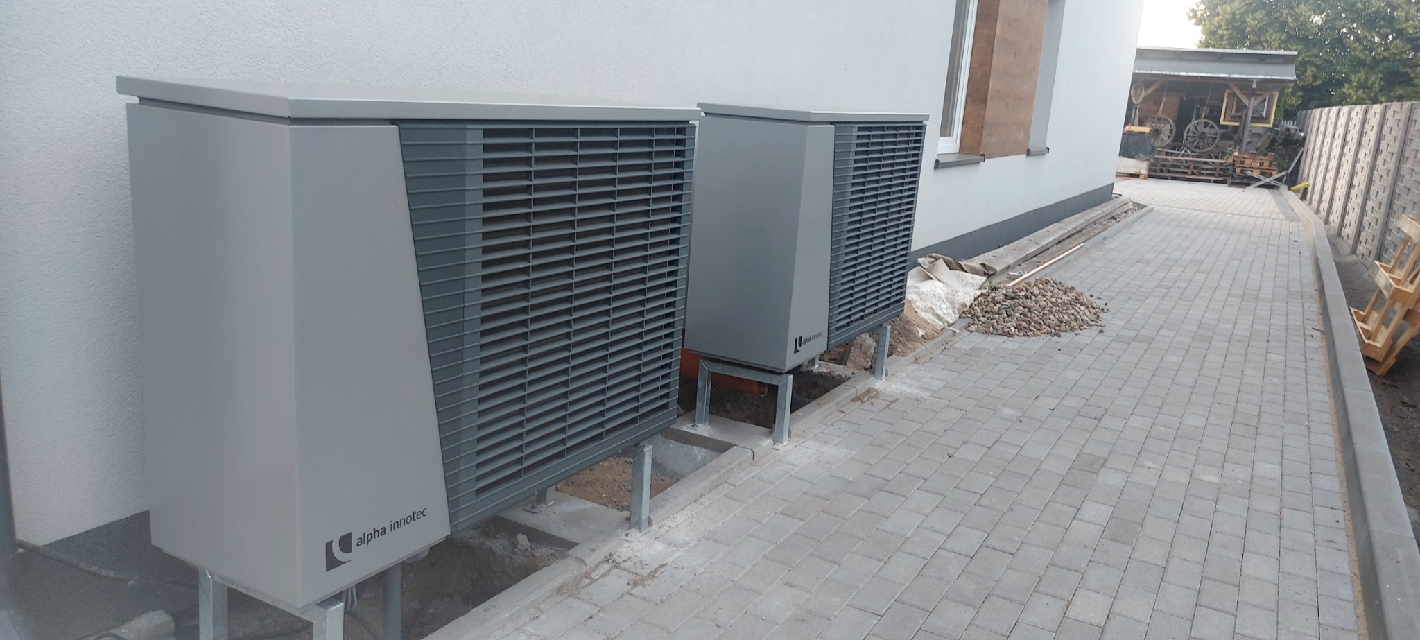 Dwie szare pompy ciepła Alpha Innotec zamontowane na metalowych wspornikach przy ścianie budynku, widoczne elementy instalacji i kostka brukowa.