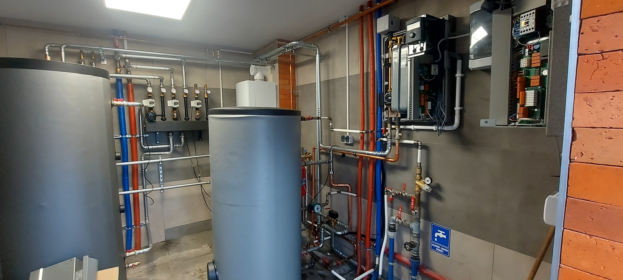 Dwa szare zbiorniki buforowe połączone rurami miedzianymi i plastikowymi (czerwone, niebieskie i białe) z zaworami i manometrami, oraz rozdzielnica elektryczna z otwartą obudową i widocznymi...