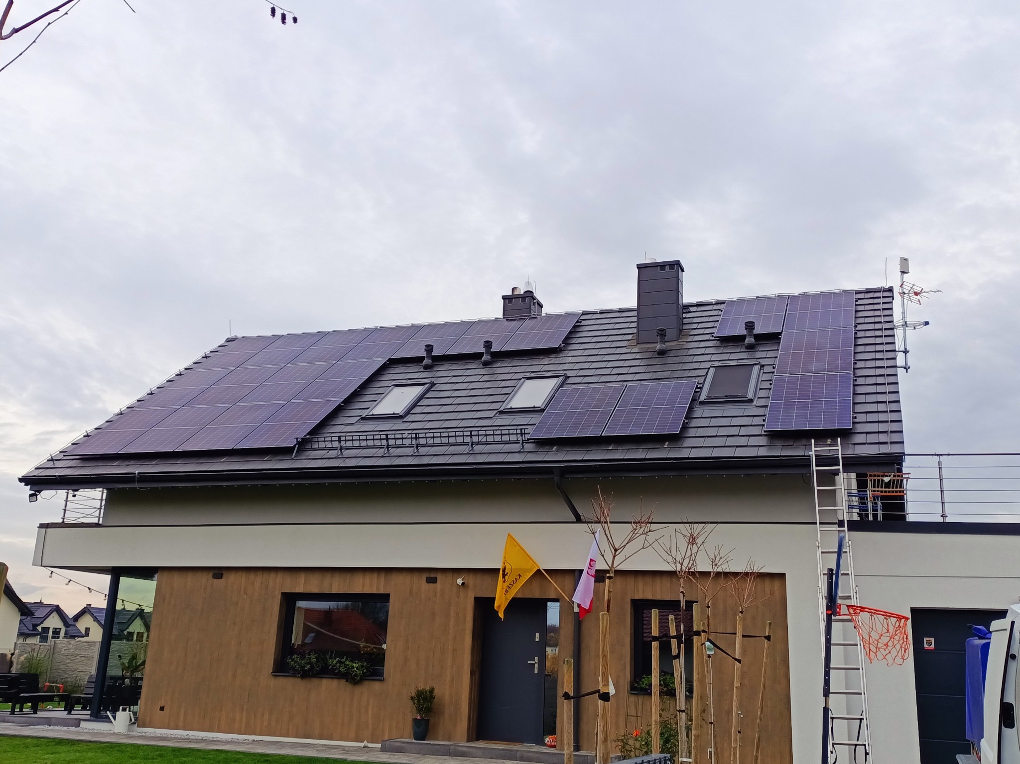 Dom jednorodzinny z zainstalowanymi panelami fotowoltaicznymi na dachu pokrytym szarą dachówką, widoczne okna dachowe i kominy, drabina oparta o ścianę, flaga i drzewka przed wejściem.
