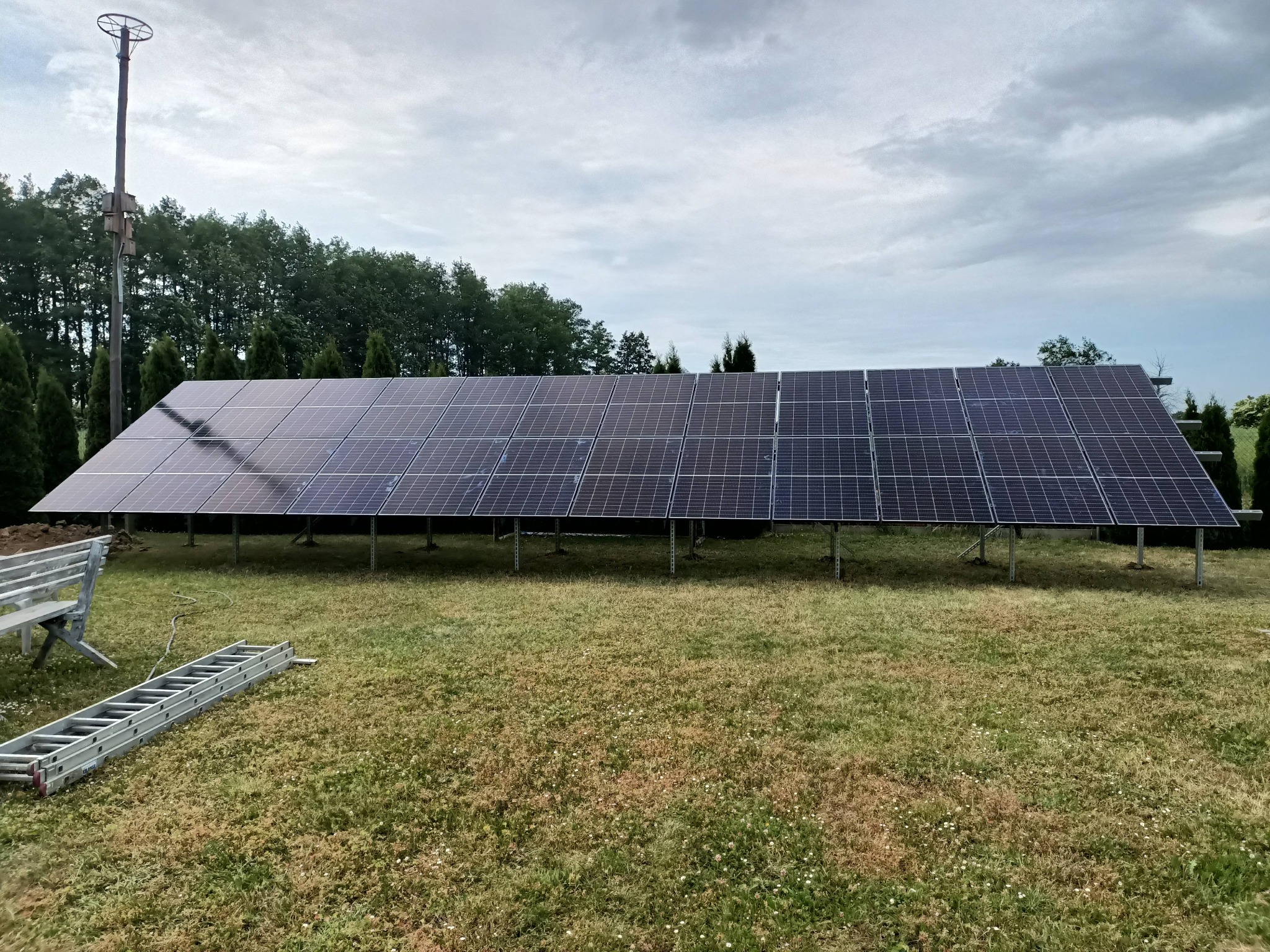 Długa instalacja fotowoltaiczna na metalowych nogach, ustawiona na trawniku przed lasem, z rozłożoną drabiną i ławką obok.