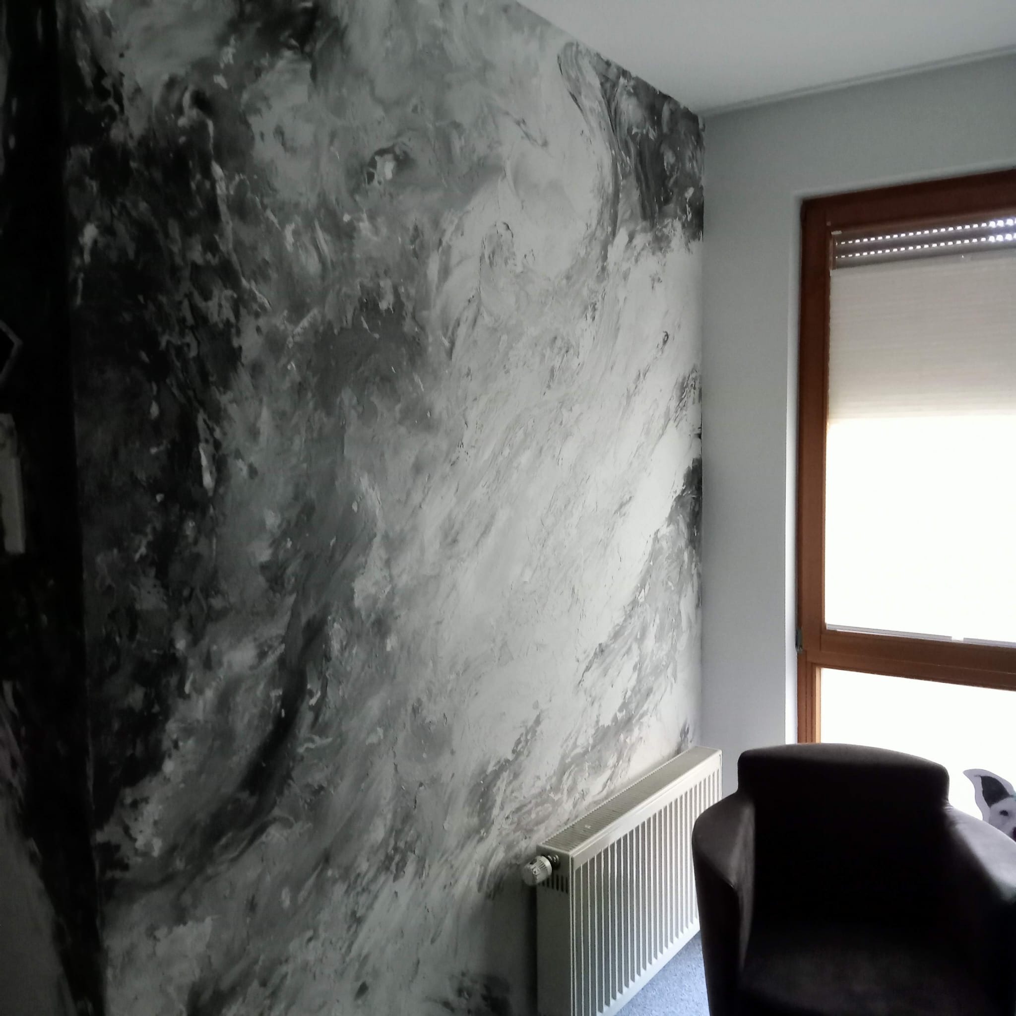 Ściana w salonie pokryta dekoracyjnym tynkiem w odcieniach szarości, przypominającym marmurkowy wzór, z widocznym grzejnikiem i fragmentem fotela.