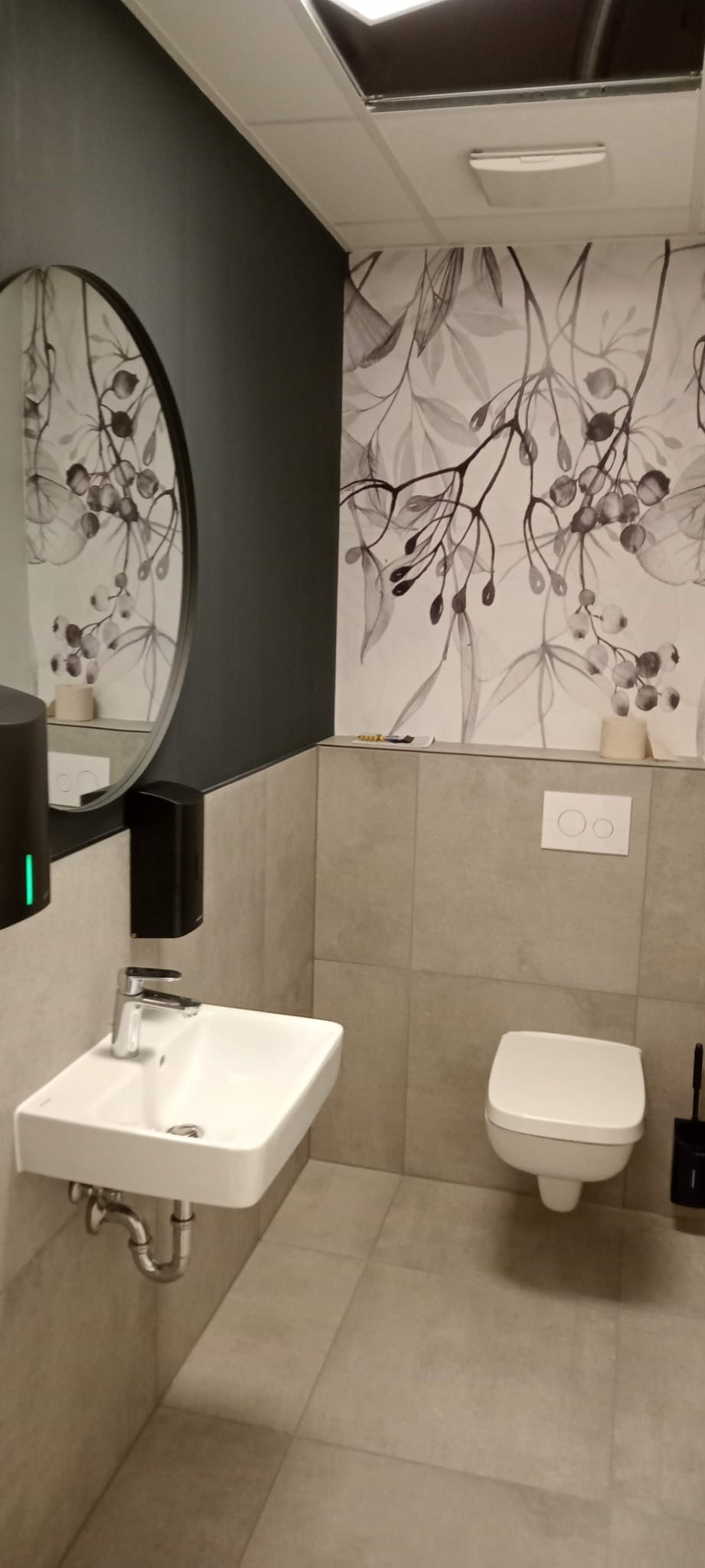 Nowoczesna łazienka z umywalką nablatową, wiszącą toaletą i tapetą w motywy roślinne, odbicie toalety w okrągłym lustrze nad umywalką, dozownik mydła z zielonym światłem.