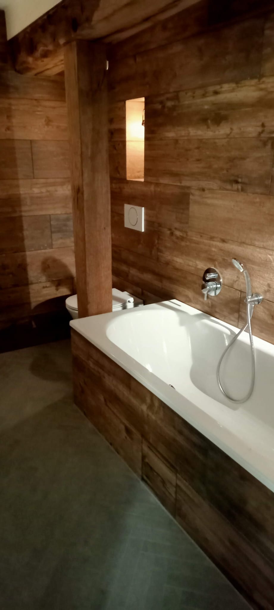 Wnętrze łazienki z drewnianą boazerią, białą wanną z chromowaną armaturą i toaletą w tle, oświetlone punktowym światłem.