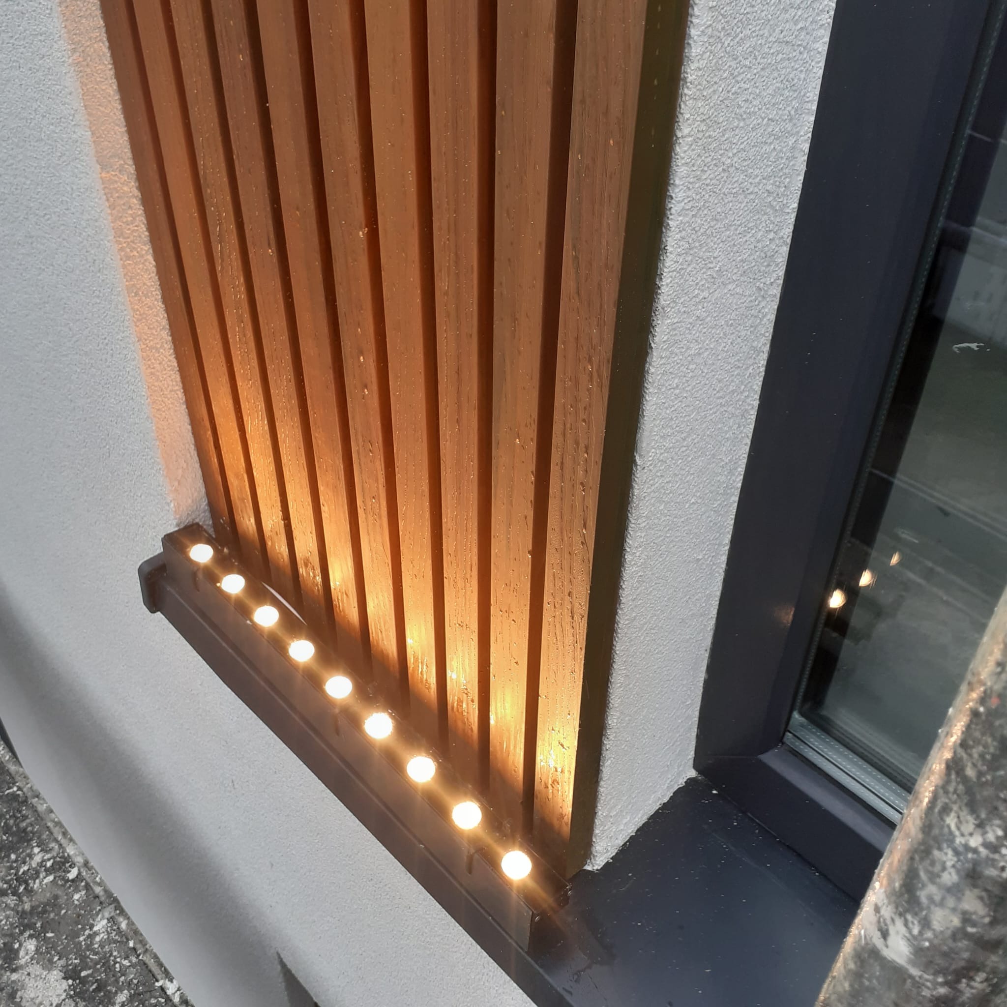 Drewniane lamele na elewacji budynku, oświetlone punktowymi lampkami LED, widok z dołu, obok okno.