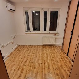 Budmik - Puste pomieszczenie po remoncie z panelami podłogowymi imitującymi drewno, białymi ścianami i oknem z grzejnikiem, widoczne instalacje elektryczne przy podłodze, klimatyzator pod sufitem.