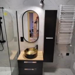 Nowoczesna łazienka z marmurowymi płytkami, czarną armaturą, złotą umywalką nablatową i owalnym lustrem z czarną ramą. W tle widoczne odbicie drabiny malarskiej.