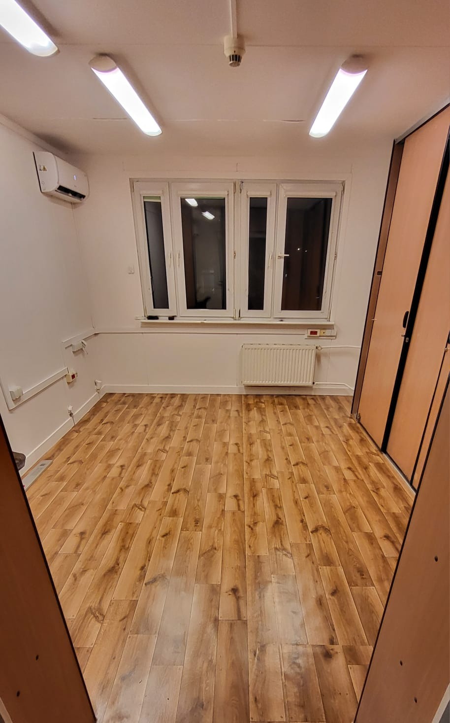 Puste pomieszczenie po remoncie z panelami podłogowymi imitującymi drewno, białymi ścianami i oknem z grzejnikiem, widoczne instalacje elektryczne przy podłodze, klimatyzator pod sufitem.