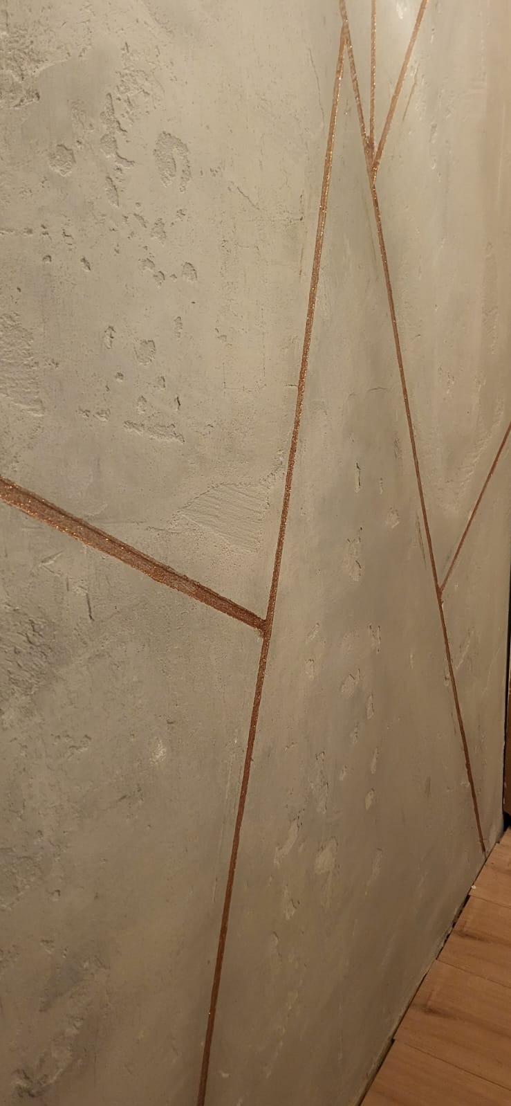 Ściana z szarą, nierówną fakturą i geometrycznymi, brokatowymi liniami w odcieniach miedzi, układającymi się w abstrakcyjny wzór.