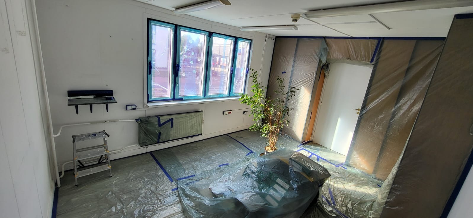 Pokój przygotowany do malowania: okna, grzejnik, podłoga i drzwi zabezpieczone folią malarską, widoczna drabina malarska i roślina doniczkowa.