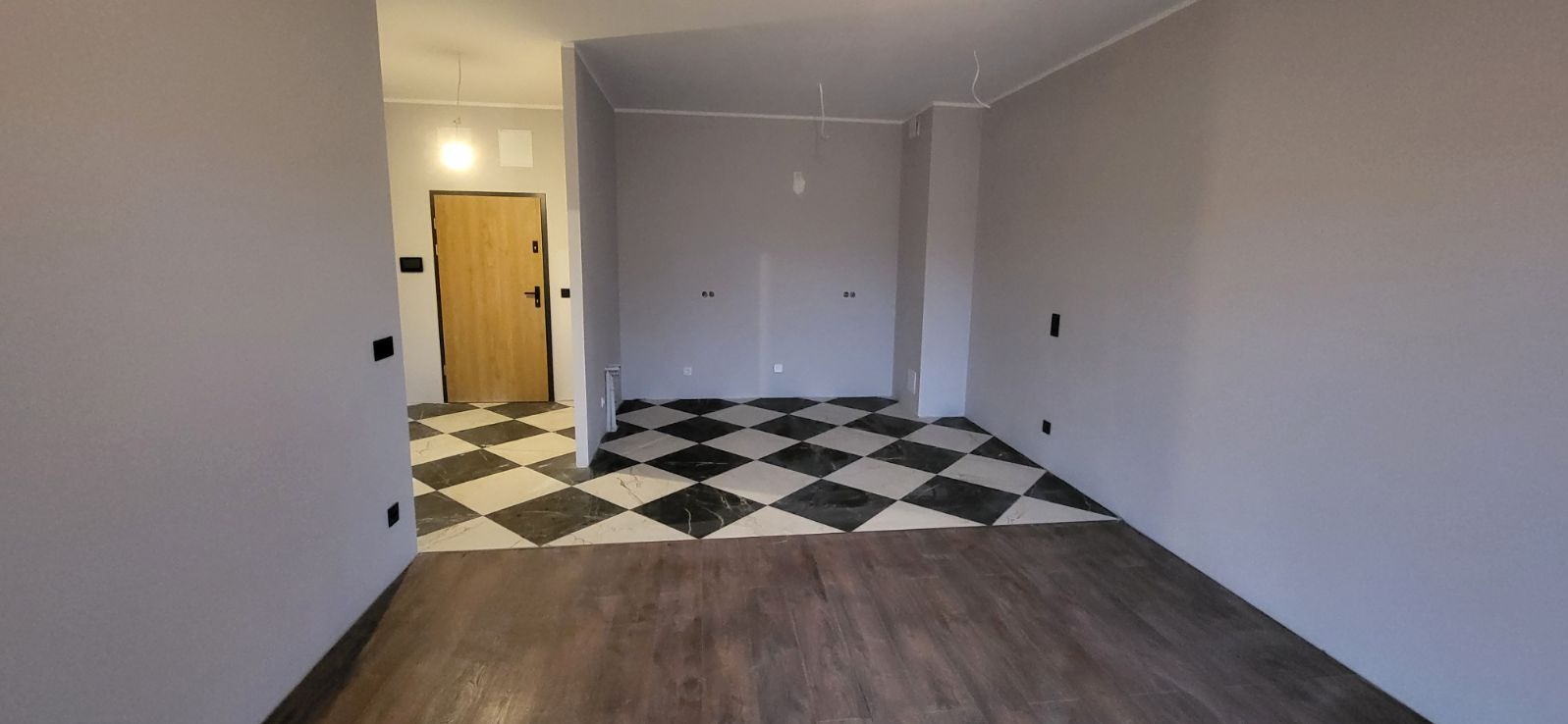 Wnętrze w trakcie wykańczania: szare ściany, podłoga imitująca drewno i czarno-biała szachownica z płytek w przejściu, widoczne niedokończone instalacje elektryczne.