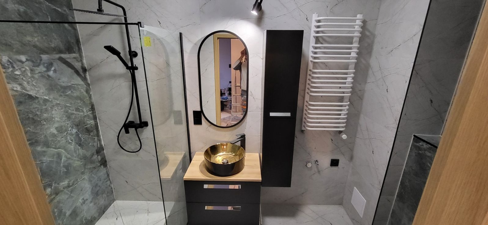 Nowoczesna łazienka z marmurowymi płytkami, czarną armaturą, złotą umywalką nablatową i owalnym lustrem z czarną ramą. W tle widoczne odbicie drabiny malarskiej.