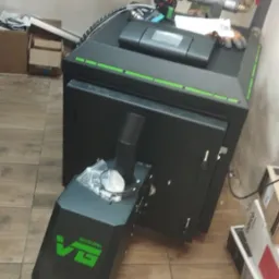 Nowoczesny piec na paliwo stałe, czarny z zielonymi akcentami, z widocznym podajnikiem i elementami instalacji grzewczej w tle, umieszczony na brązowych płytkach podłogowych.