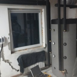 Hydro-dom - Szary zasobnik ciepłej wody użytkowej z czarnymi rurami instalacji grzewczej w trakcie montażu, widoczna drabina, walizka z narzędziami i miedziane rury.