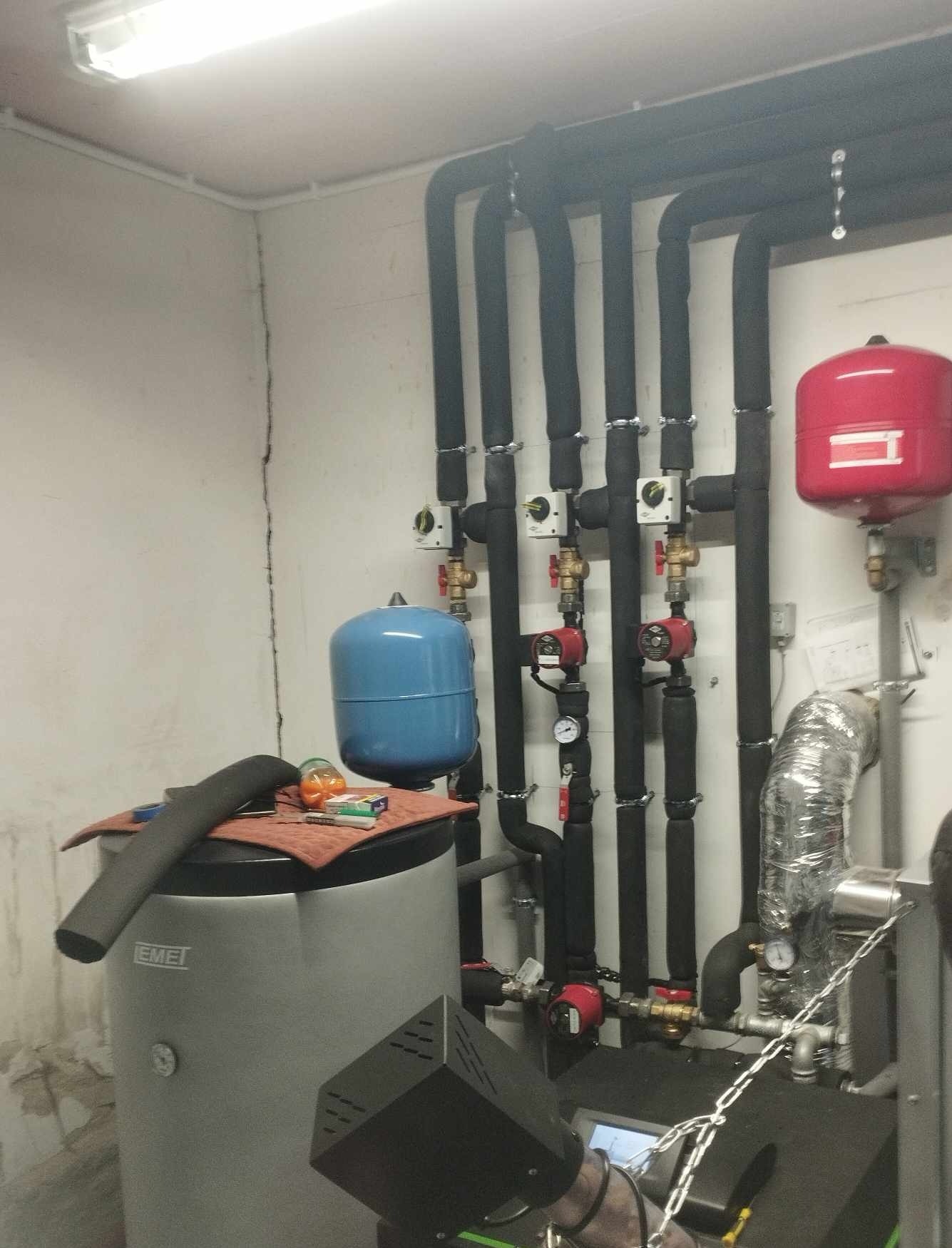 Kotłownia z szarym bojlerem, czarnymi rurami z izolacją, czerwonym i niebieskim zbiornikiem wyrównawczym. Na bojlerze leżą narzędzia i materiały instalacyjne.