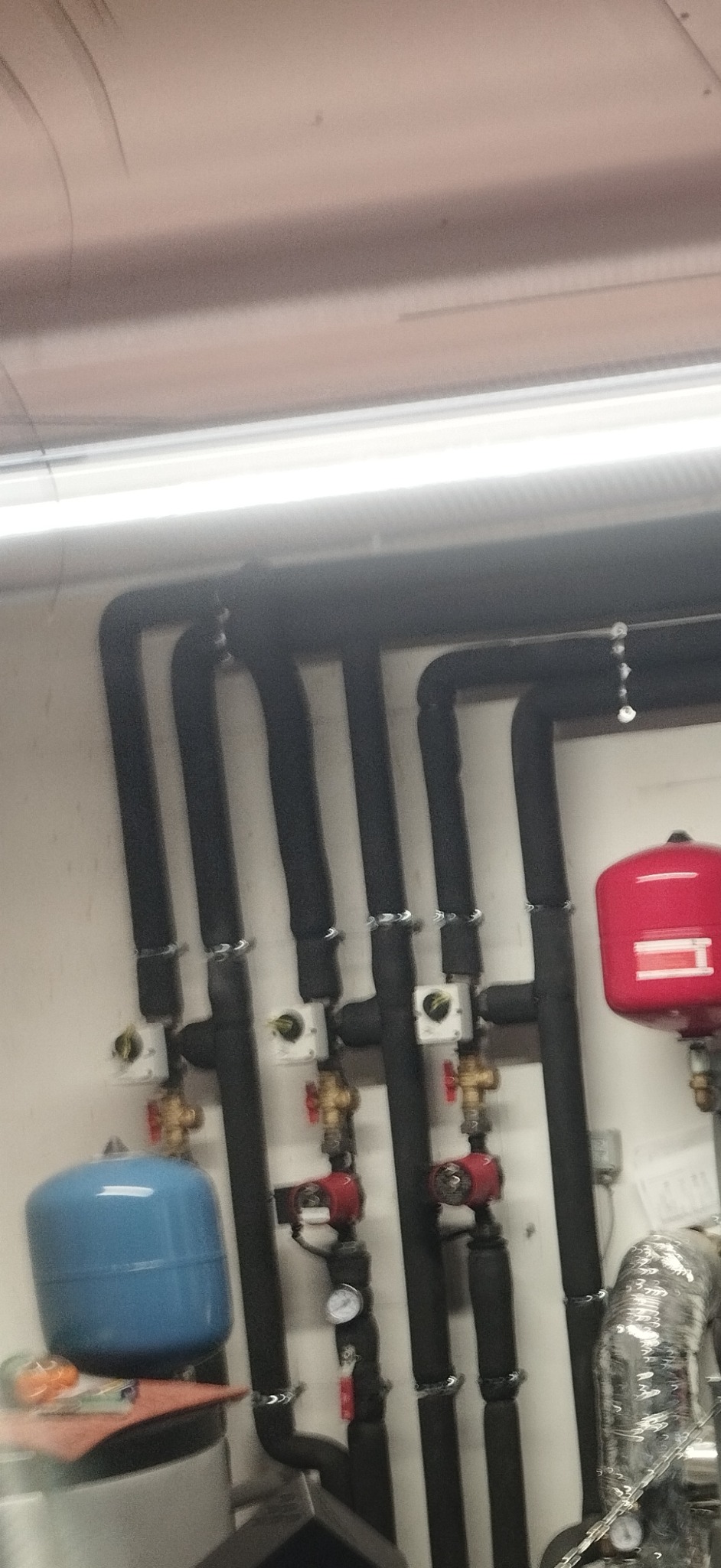 Szereg czarnych, zaizolowanych rur instalacji grzewczej z zaworami i manometrami, prowadzonych wzdłuż ściany, z widocznymi zbiornikami wyrównawczymi w kolorach niebieskim i czerwonym.