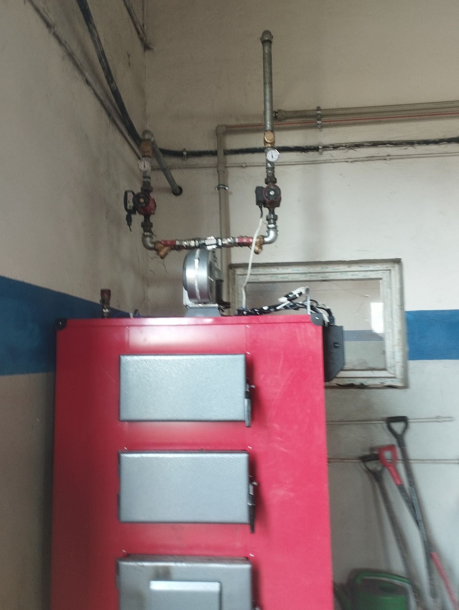 Czerwony piec z trzema szarymi drzwiczkami, podłączony do instalacji rur z manometrami, na tle ściany z niebieskim paskiem i narzędziami.