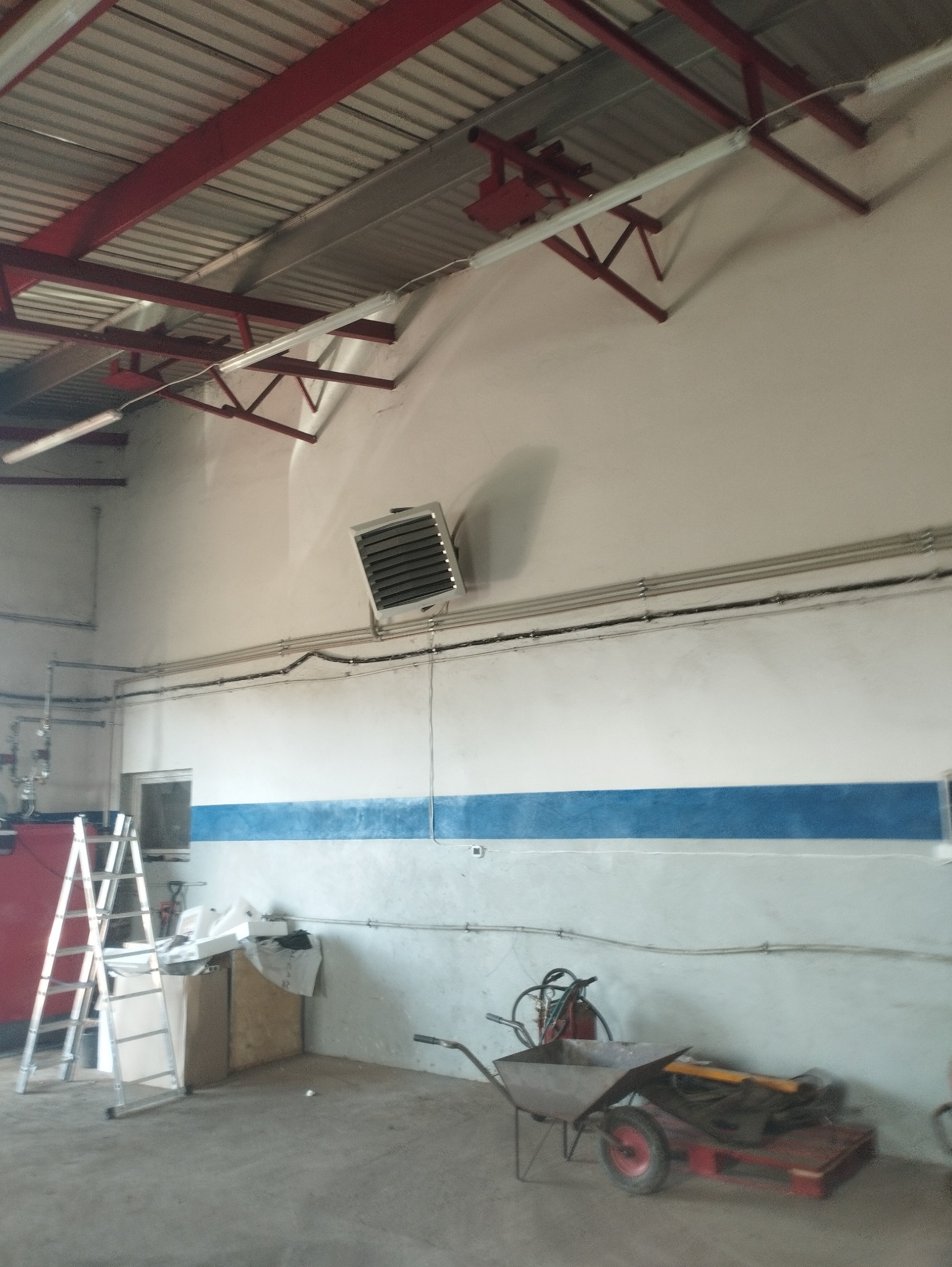 Nagrzewnica przemysłowa zamontowana na ścianie w hali, widoczne rury instalacyjne, drabina i taczka z narzędziami w tle.