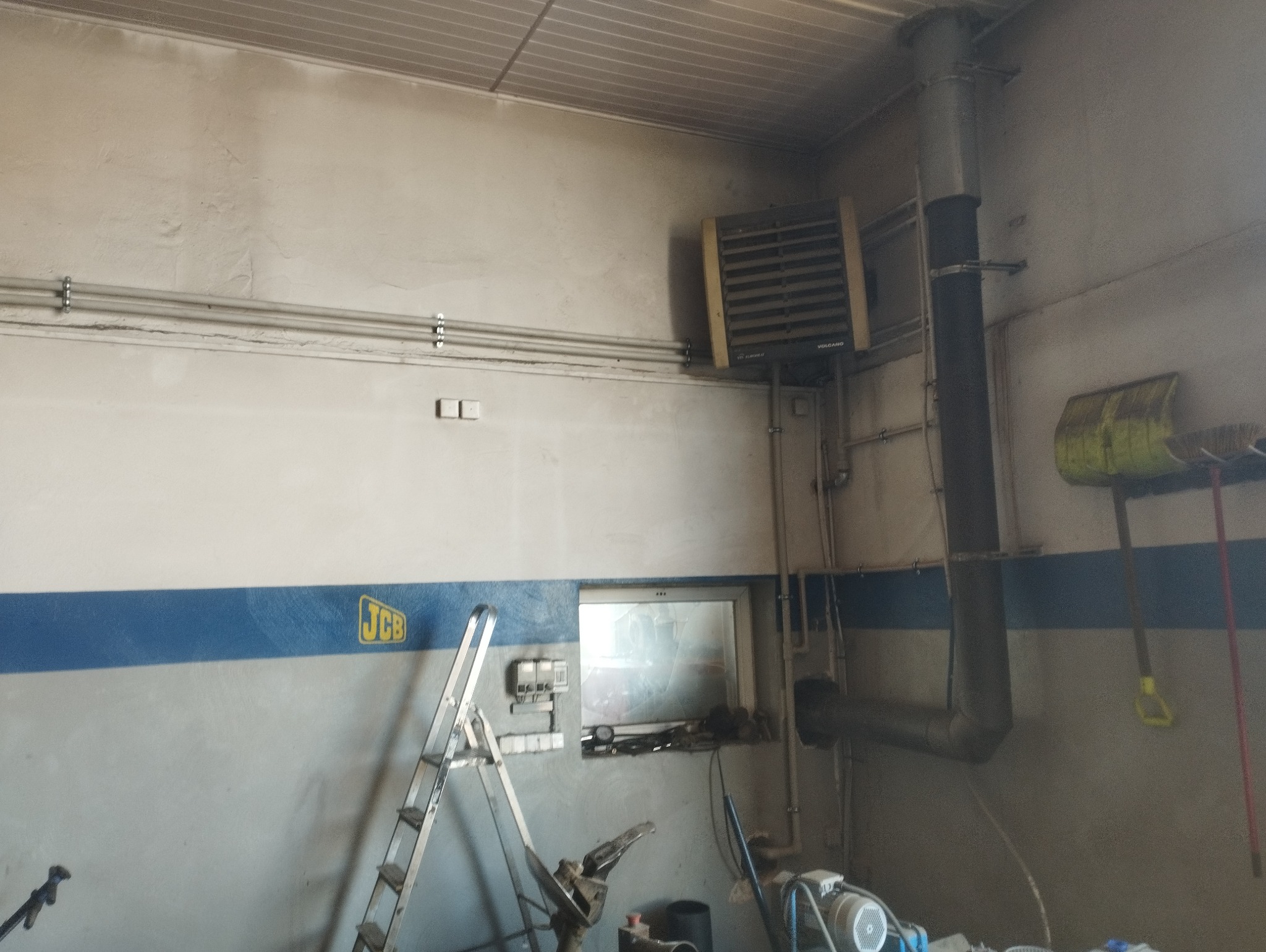 Wnętrze warsztatu z zamontowanym nagrzewnicą Volcano z szarą rurą odprowadzającą spaliny, drabina aluminiowa oparta o ścianę z niebieskim pasem i logiem JCB, widoczne narzędzia i sprzęt warsztatowy.