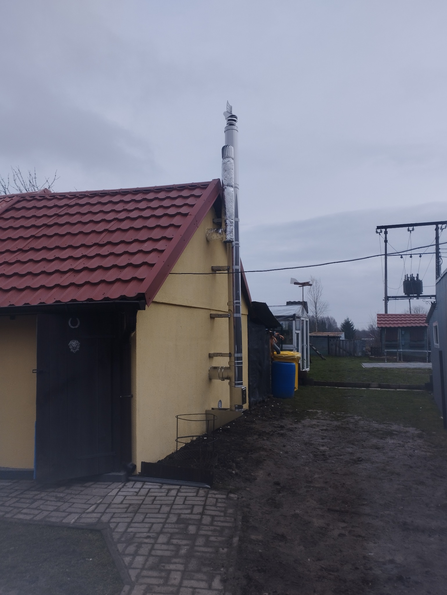 Zewnętrzny komin systemowy ze stali nierdzewnej, zamontowany na elewacji budynku z czerwoną dachówką, z widocznymi elementami mocującymi i izolacją termiczną, na tle pochmurnego nieba.