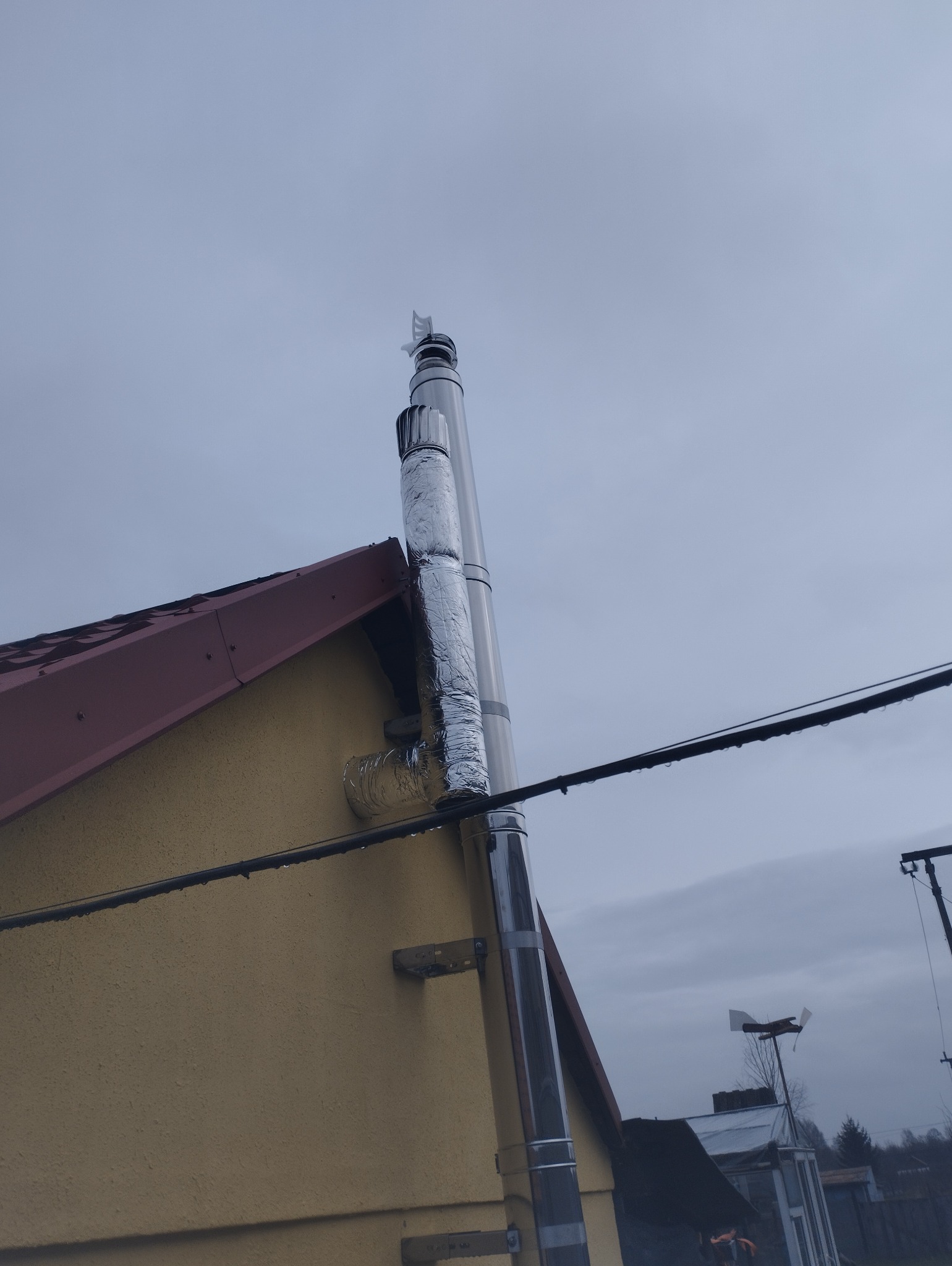 Zewnętrzny, ocieplony komin spalinowy ze stali nierdzewnej, zamontowany przy ścianie budynku z żółtą elewacją i czerwoną dachówką, widok z dołu na pochmurne niebo.